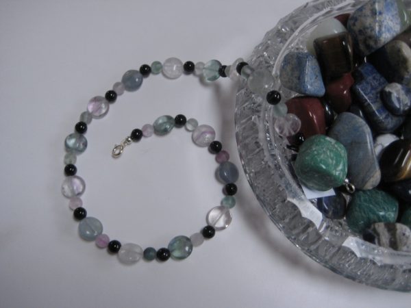 Edelsteinkette-aus-Fluorit-und-Turmalin, aus einer Glasschale mit Edelsteinen fließend. Im Bild ist auch ein Türkis in der Schale.