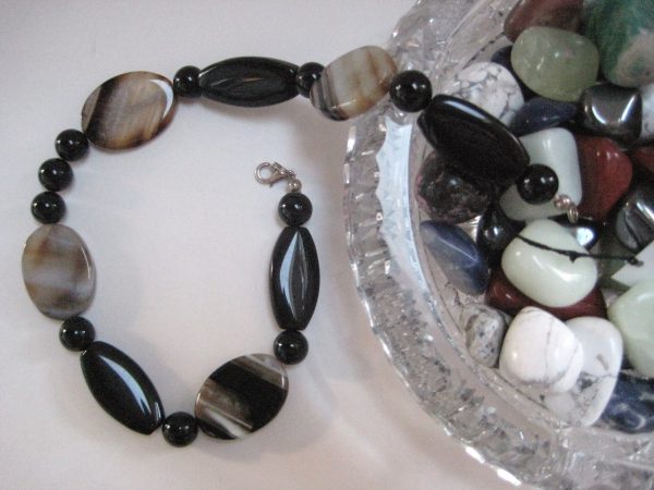 Eine Edelsteinkette aus Achat und Onyx mit Steinen in Grau- und Schwarztönen fließt aus einer Glasschale mit anderen Edelsteinen.