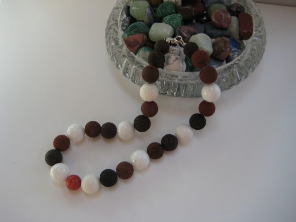 Eine Edelsteinkette aus Lavastein, Jade und Koralle in schwarz, weiß und braun fließt aus einer Glasschatulle.