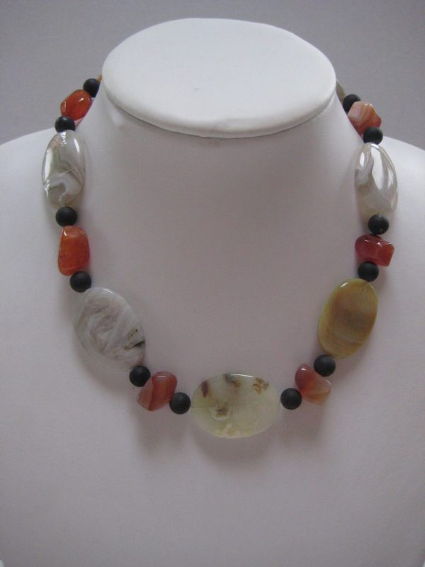 Eine Edelsteinkette aus Onyx, Achat und Karneol auf einer weißen Damenbüste. Sie besteht aus Achat-Elipsen in verschiedenen Farbtönen mit Onyxkugeln und orangen Karneolnuggets.