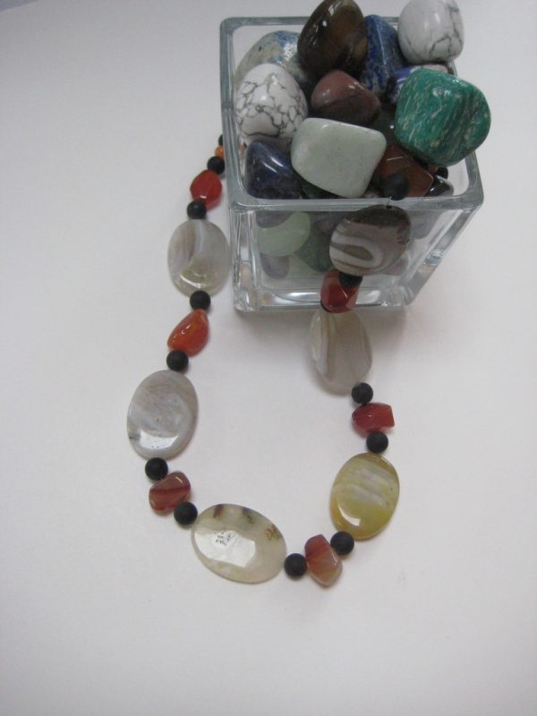 Eine Edelsteinkette aus Onyx, Achat und Karneol, die aus einem Glasbehälter mit Edelsteinen fließt. Sie besteht aus Achat-Elipsen in verschiedenen Farbtönen mit Onyxkugeln und orangen Karneolnuggets.