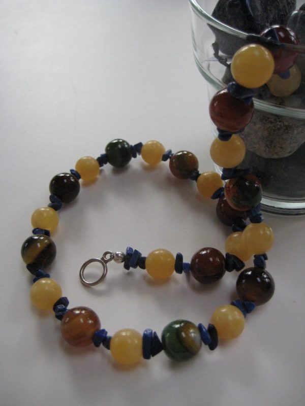 Eine Edelsteinkette aus Orangencalzit, Lapislazuli und Achat fließt aus einer Glasschale. Die Farben reichen von Gelb bis hin zu Brauntönen.
